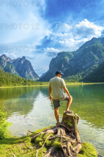 Tourist standing on a tree stump admires the breathtaking landscape of lake dobbiaco, surrounded by the majestic dolomites on a sunny summer day