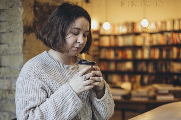 A woman in a soft sweater holds a takeaway coffee cup while enjoying a peaceful moment in a cozy bookstore Bookshelves in the background create a warm and relaxed atmosphere