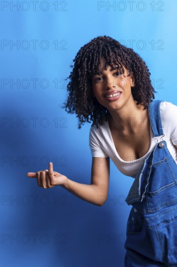 Young woman with curly hair pointing her finger to the left side of the frame while smiling on a vibrant blue background