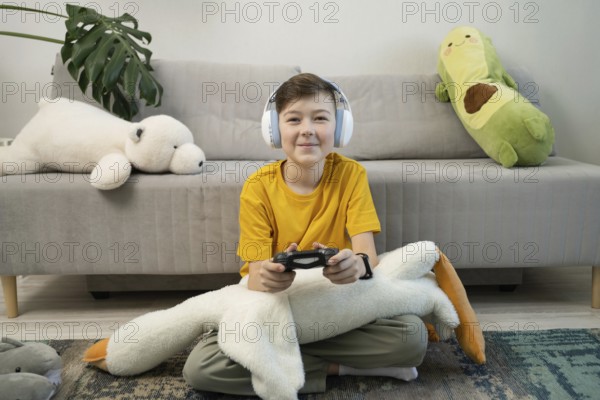 A young boy in a yellow shirt plays video games while sitting cross-legged on a rug. Surrounded by stuffed animals, he wears headphones, embracing a cozy, playful atmosphere