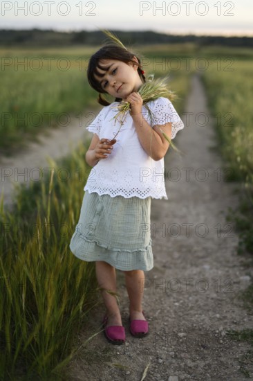 A young girl, identifiable by her feminine dress and hairstyle, stands on a dirt path with wheat spikes in hand, set against a beautiful rural backdrop. She appears contemplative and connected to the natural environment around her