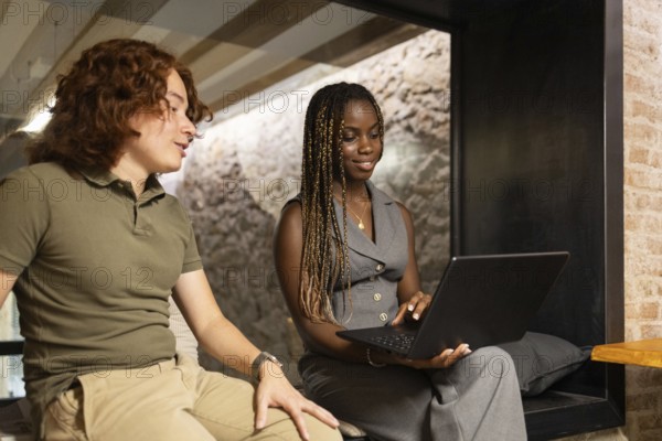 Two young professionals an African woman and a caucasian red hair man engaged in a productive coworking session. A woman holds a laptop while discussing an innovative idea, highlighting a collegial office environment