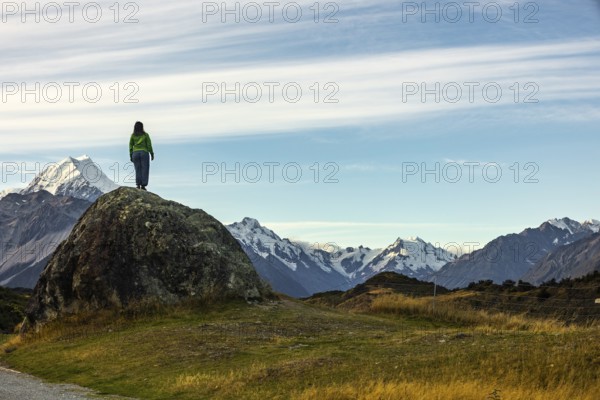 A woman stands on a rocky outcrop overlooking Mount Cook in New Zealand, surrounded by stunning autumn scenery. The snow capped peaks and clear skies create a breathtaking backdrop
