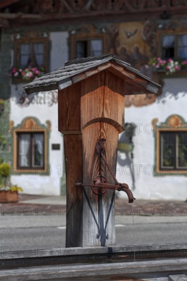 A traditional wooden fountain in front of a Bavarian building with decorative flowers and sunshine, Wallgau, Bavaria, Germany