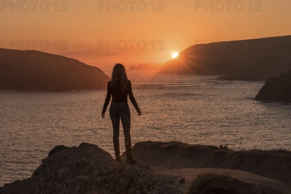 A silhouette of a woman stands on a rocky cliff at Arnia Beach, Costa Quebrada, gazing toward the glowing sunrise over the Atlantic coastline in northern Spain