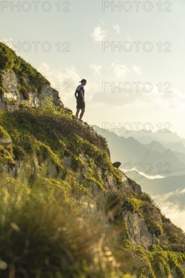 A lone man stands on a scenic mountain trail, taking a moment to rest during an invigorating run. The serene landscape surrounds him with lush greenery and distant peaks