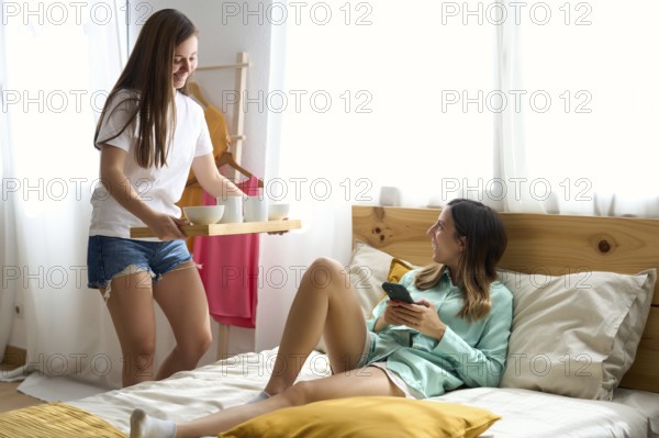 A joyful lesbian couple shares a tender moment in their sunlit bedroom One woman serves breakfast while the other relaxes with her phone, celebrating love and togetherness