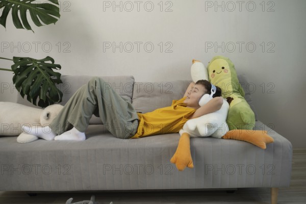 A young boy in a yellow shirt lies comfortably on a grey sofa, wearing headphones and surrounded by plush toys, under a large green plant, enjoying a moment of relaxation