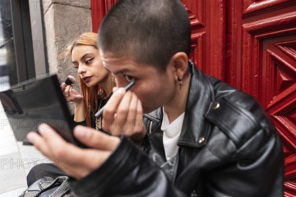 A lesbian couple sits together in front of a red door, each applying makeup with focus and care, highlighting a sense of individuality and shared moments