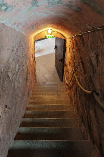 A dimly lit stone tunnel with a staircase and a rope railing leading to an open door area, Le Mont-Saint-Michel
