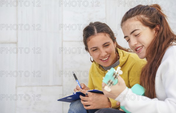 Two friends engage in a fun study session One plays a turquoise ukulele, while the other takes notes on a clipboard Both are smiling, enjoying the shared musical moment
