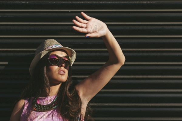 Woman wearing a summer hat and sunglasses raises hand to shield eyes from the sun. The background features a dark, striped wall, adding contrast to the scene