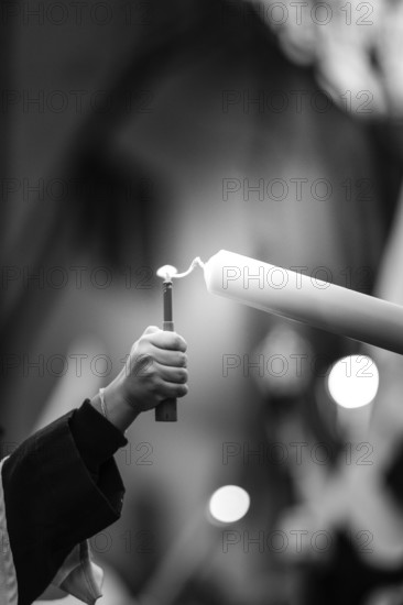 A monochrome image depicting a hand holding a lighter, reaching to ignite a tall candle, symbolizing light and spirituality in holy week. The blurred background enhances the focus on the flame