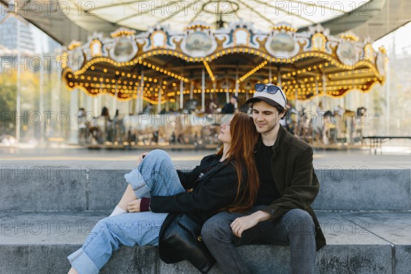 A young couple sits on steps in front of a brightly lit carousel in Brooklyn Bridge Park. They exude relaxation and happiness, enjoying a sunny day in an urban setting, surrounded by street activity