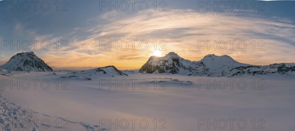 The sun sets behind the rugged, snow-covered peaks of Katla Glacier, casting a golden glow over the vast, untouched winter landscape. A dramatic sky completes this breathtaking panoramic view