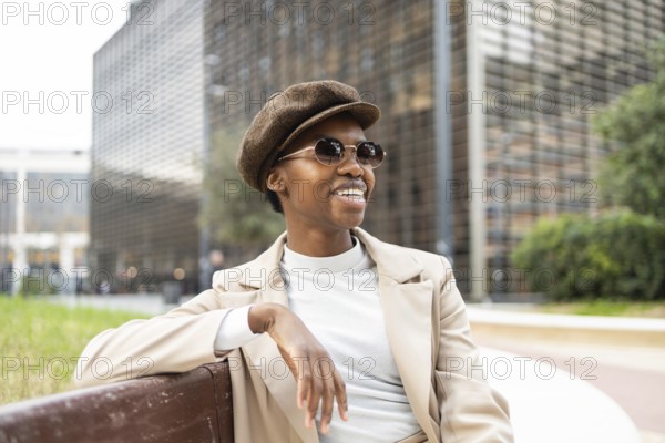 A fashionable black woman in sunglasses and a hat sits on a city bench, exuding confidence and style in an urban environment, surrounded by modern architecture