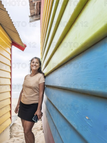 A smiling woman leans against the colorful Brighton Bathing Boxes in Melbourne, Australia. The vibrant hues of the iconic beach sheds create a lively and cheerful atmosphere