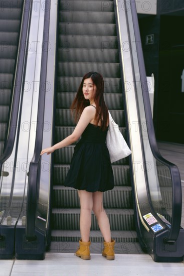 A young asian woman in a black dress and boots stands on an escalator in an urban setting, holding a tote bag. The scene captures a modern city lifestyle