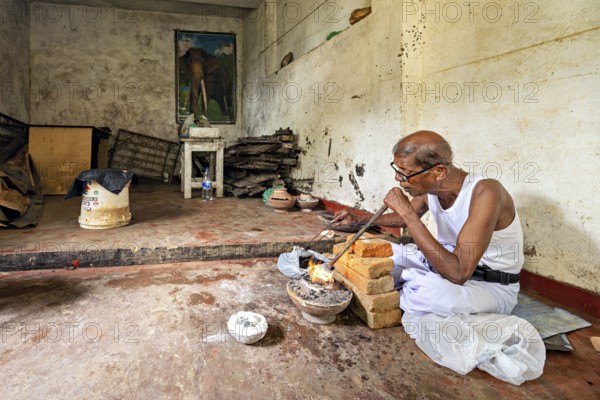 An old man works silently in a workshop with bricks and fire, an elephant poster on the wall, a man burns sapphires in a small oven in the town of Ratnapura in Sri Lanka