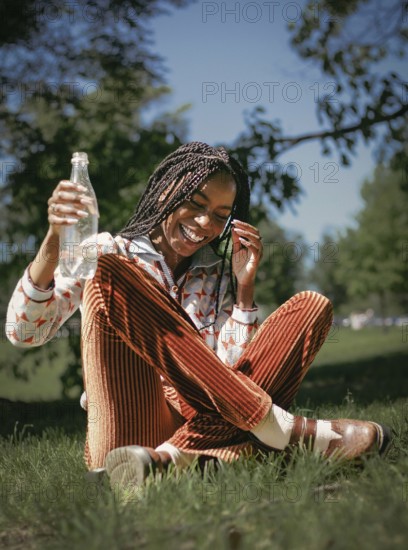 A joyful African American woman with braided hair laughs heartily while sitting on grass and holding a water bottle. The sun shines brightly, adding warmth to the serene scene