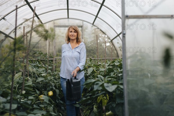 A woman stands in a lush greenhouse, holding a watering can. Surrounded by thriving plants, she embodies sustainable agriculture and care for the environment