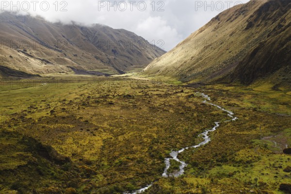 A meandering stream flows through a vast Andean valley at El Altar volcano, Ecuador, surrounded by rolling hills, golden grasses, and dramatic light and shadow contrasts