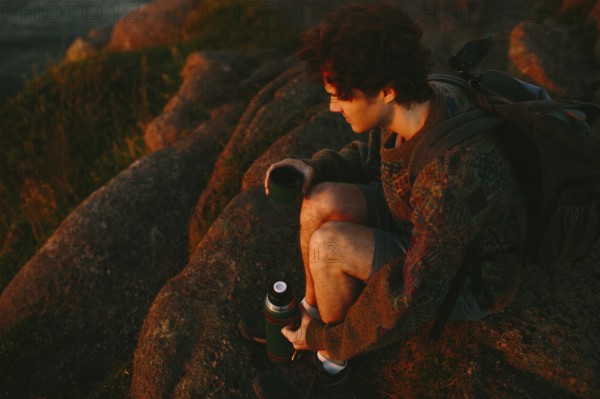 A young male hiker sits on rugged rocks at Cape Spear Lighthouse, watching the sunset. He is holding a thermos and relaxing after a day of hiking