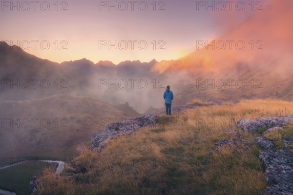 A lone person stands on a grassy ridge, gazing at misty mountains during dusk in summer. Warm light bathes the landscape, evoking a sense of peace and tranquility in the Pyrenees