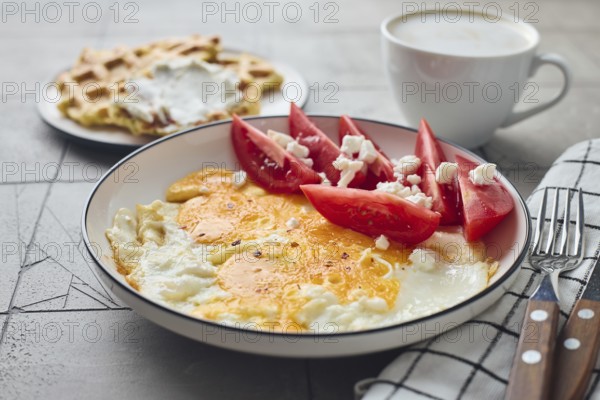 From above a delicious homemade breakfast featuring fried eggs with feta cheese and fresh tomato slices alongside Belgian waffles topped with cream cheese and a steaming cup of coffee