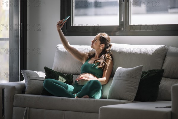 Pregnant woman sitting comfortably on a couch, wearing a yoga outfit, and taking a selfie. She embraces wellness and calm in a peaceful home environment, enjoying a serene moment