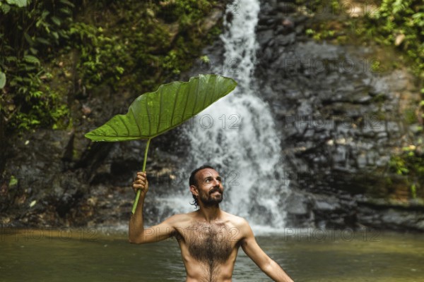 Shirtless man holding a large leaf smiles as he uses it for shade while standing in a water pool in front of a waterfall in the lush Choco Andino rainforest of Ecuador