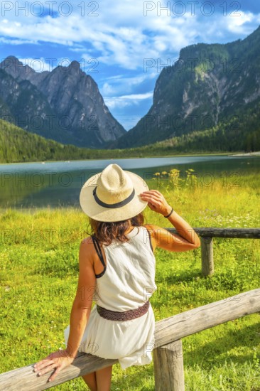 Woman wearing straw hat admiring lake dobbiaco with the dolomites in the background on a sunny summer day