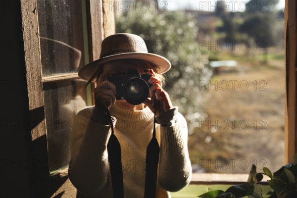 In a cozy countryside home near Barcelona, Spain, a senior unrecognizable woman captures the serene landscape using an analog camera. She is dressed in a warm sweater and stylish hat, deeply engaged in her photography