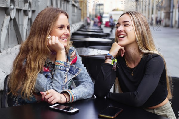 Two teenage girls smile and chat outdoors, with cell phones on the table, enjoying each other's company and the moment of connection in a lively urban setting