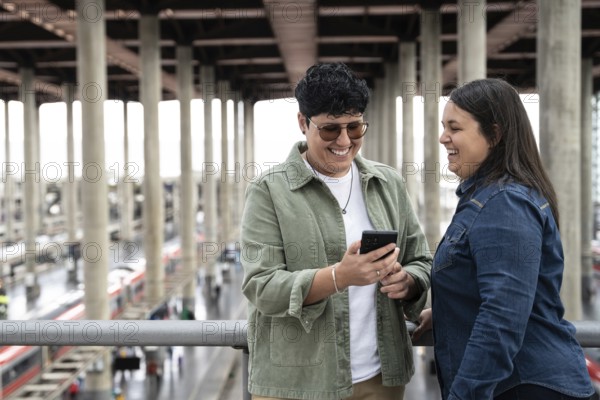 A joyful moment between a lesbian curvy couple at a train station, sharing smiles and a phone, embodying love and companionship in a travel setting