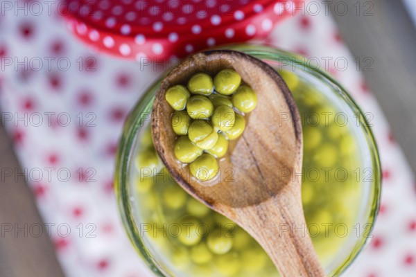 A wooden spoon holds green homemade organic peas over a glass jar, set on a white cloth with red polka dots. The vibrant colors and textures create a fresh, inviting scene