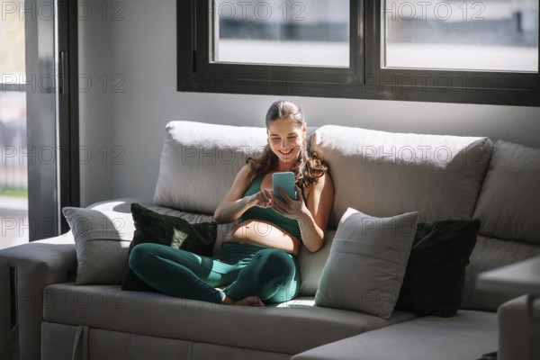 A pregnant woman sits comfortably on a cozy sofa, wearing a yoga outfit. She enjoys her leisure time, focusing on wellness and connecting digitally through her smartphone