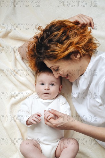 A mother shares a tender moment with her newborn baby girl, lying on a soft, cream-colored blanket. The baby wears a white onesie, creating a serene and loving atmosphere