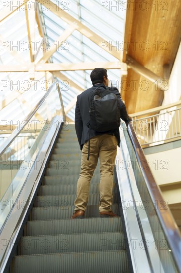 Young Chilean businessman in smart attire with a backpack ascends an escalator in a modern, brightly lit building. The scene conveys ambition and upward mobility