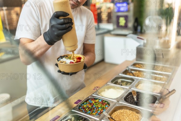 Barista wearing black gloves preparing a customized acai bowl, adding a creamy sauce over fresh fruits, granola, and other various toppings in a paper cup in a modern cafe setting