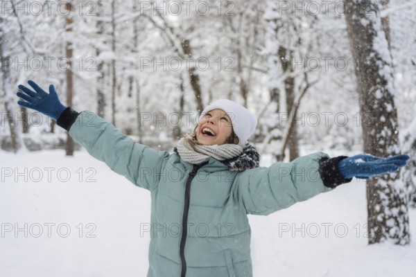 A cheerful child in a blue jacket and white hat enjoys a snowy day in a winter forest, embracing the cold with a big smile. Snowflakes gently fall around them, creating a serene scene