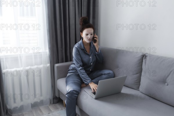 A woman in pajamas and a facial mask sits on a gray couch, multitasking between a laptop and a phone. The room features soft natural light through a window