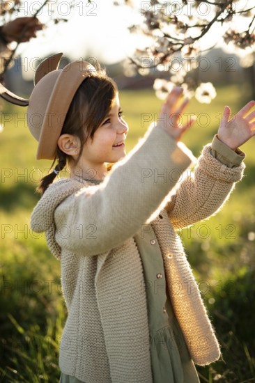 A joyful little girl in a beige hat and sweater plays under the almond blossoms on a sunny spring day, expressing happiness and curiosity