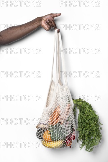 A hand holds a reusable cotton mesh bag filled with fresh vegetables and fruits, showcasing sustainable shopping practices against a clean white background, promoting eco-friendly habits