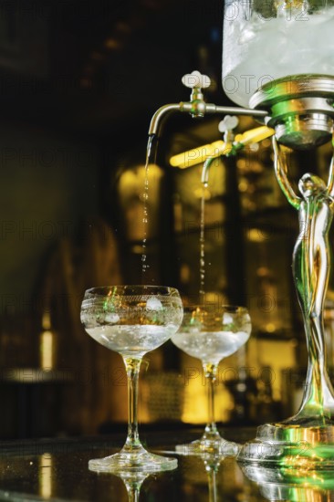 Elegant setup with absinthe fountain, gently dripping ice water into vintage crystal glasses. The ambient lighting creates an inviting atmosphere for this classic ritual