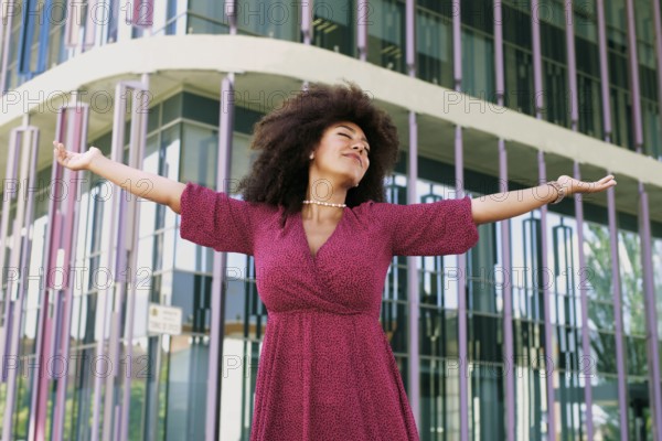 Young Arab woman with afro hair, wearing a pink dress, joyfully extends her arms in front of a modern glass building with eyes closed. She exudes confidence and empowerment