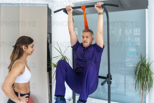 Physiotherapist observing a man performing leg raises on a pull up bar, engaging in medical recovery exercises or sports osteopathy therapy at a clinic