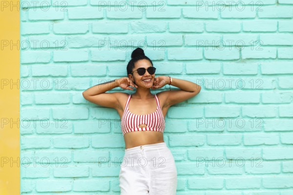 A young Latin woman stands against a vibrant turquoise wall in Mancora, Peru. Wearing a striped top and sunglasses, she exudes joy and summer vibes in this lively beach town