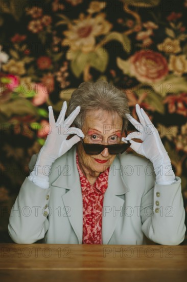 Elegant elderly woman posing confidently, wearing white gloves and sunglasses. Her stylish outfit stands out against a floral studio background, exuding grace and charm