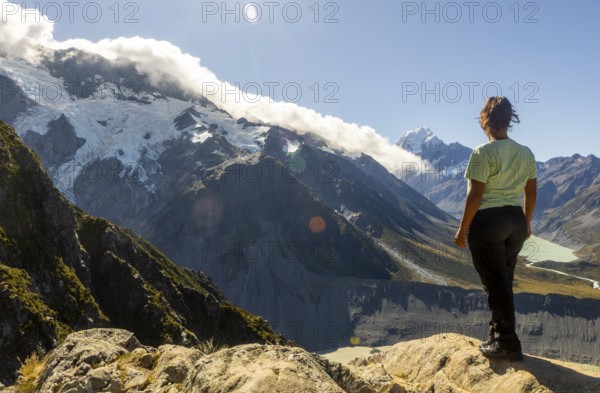 A woman enjoys the stunning autumn view of Mount Cook, New Zealand, standing on a rocky vantage point. The scene captures the majestic natural beauty of the region